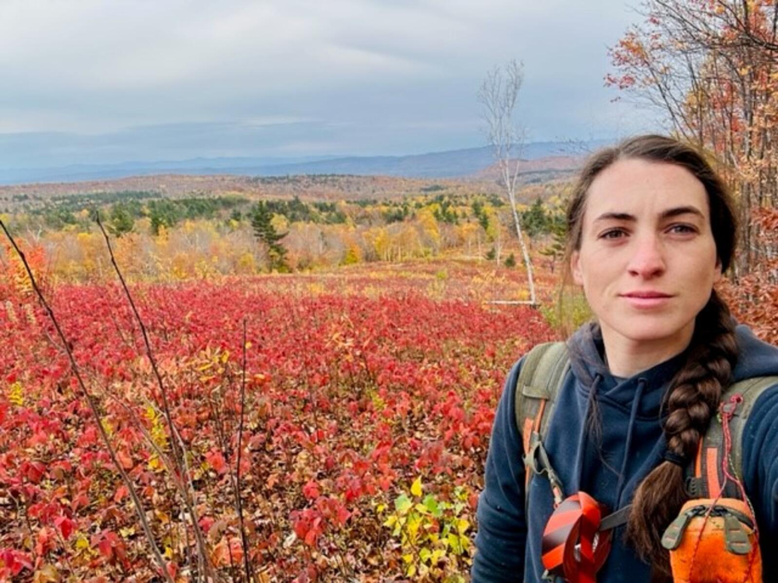 Selfie of Claire with long dark side braid and orange red fall color landscape of trees in background