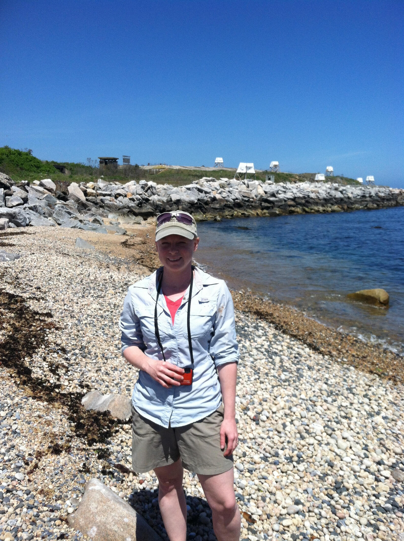 Victoria stands on a small waterfront beach area with many rocks and shells, wearing a blue button down holding binoculars