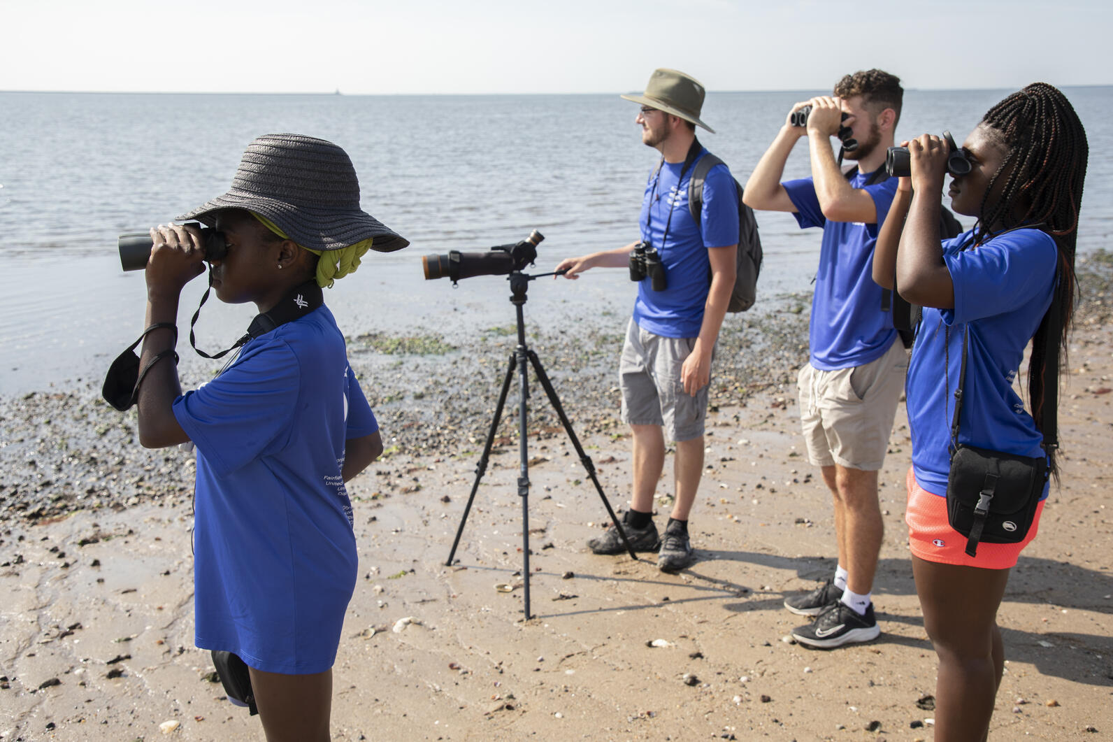 A group of young adults in matching blue work shirts stand on the beach, using binoculars to look at something in the distance towards the shoreline.