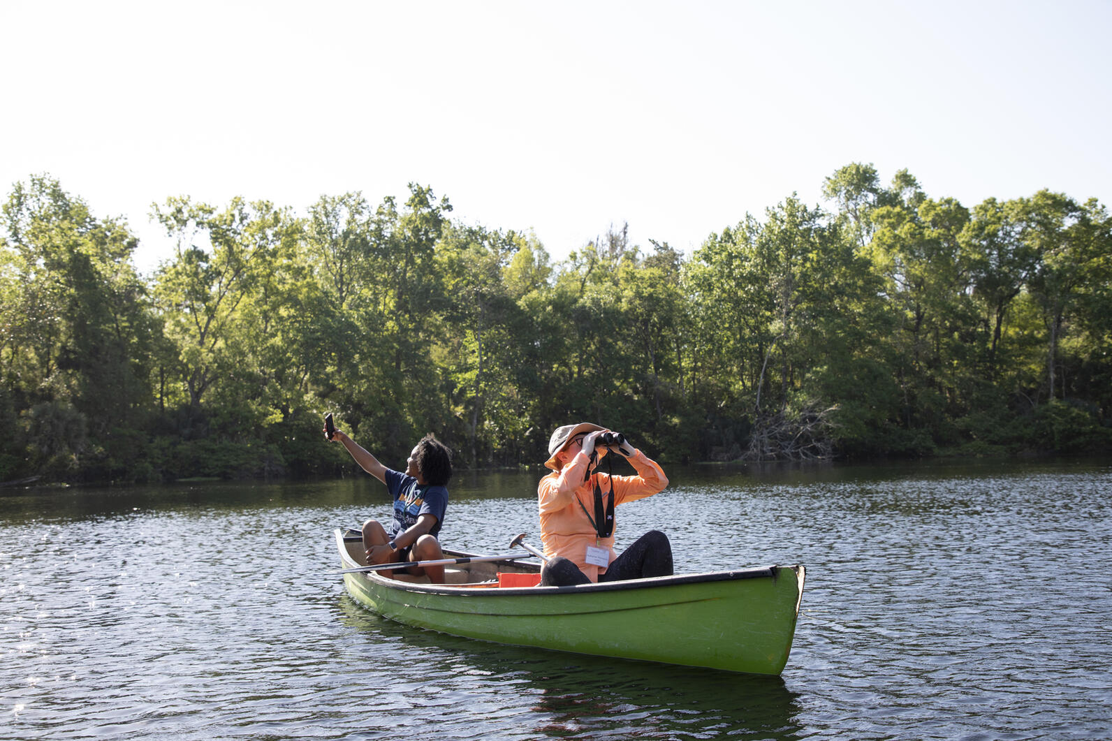 2 young people in a kayak on a lake. The person in the front is using binoculars while the person in the back is taking a selfie. 