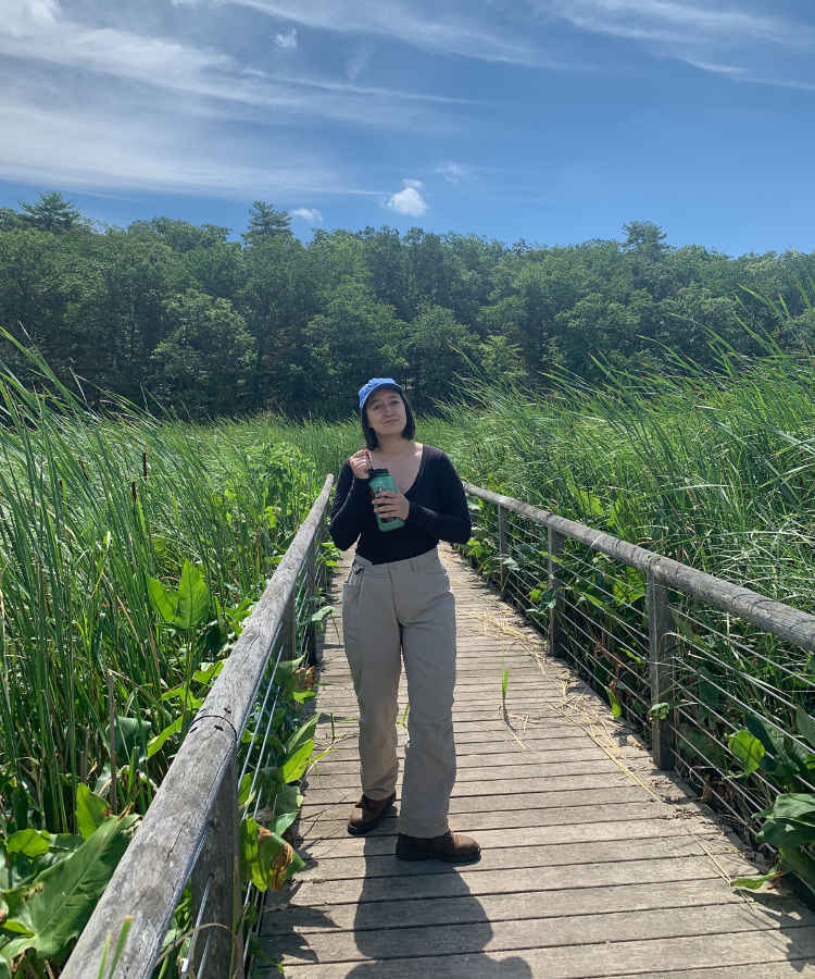 A person in a black shirt and long khaki-colored pants stands on a wooden boardwalk on a sunny day. On either side of the boardwalk are green marsh plants.