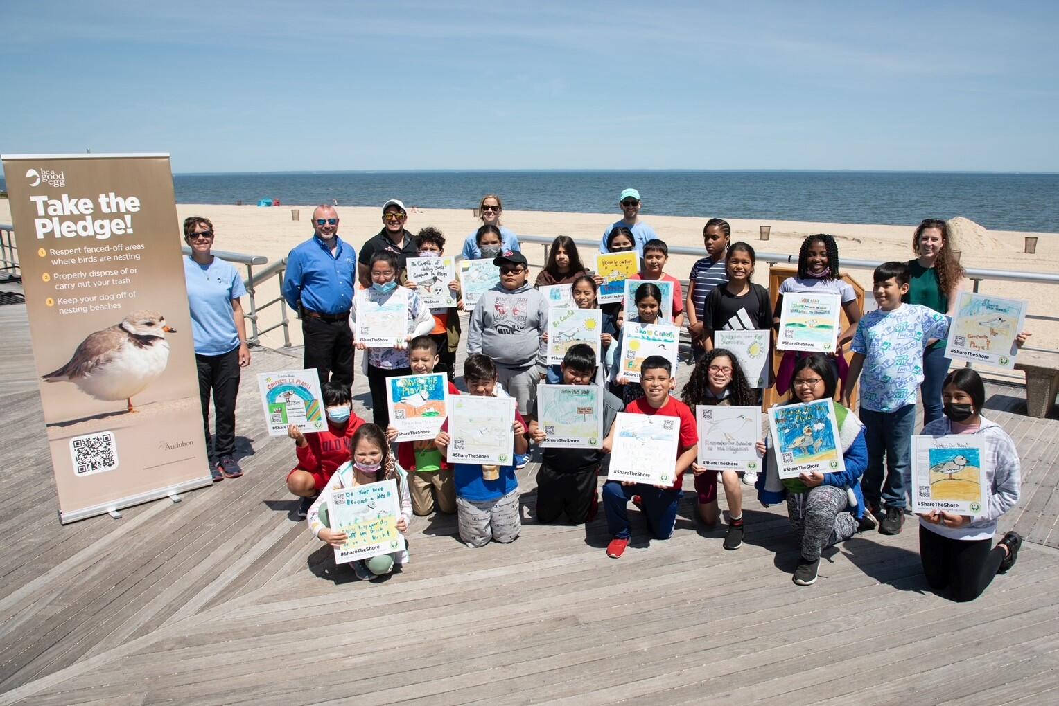 A large group of children with several adults smiling at a camera on a beach boardwalk. Each child holds a sign showing a hand-drawn illustration warning people to stay out of shorebird nesting sites, drawn by the children.