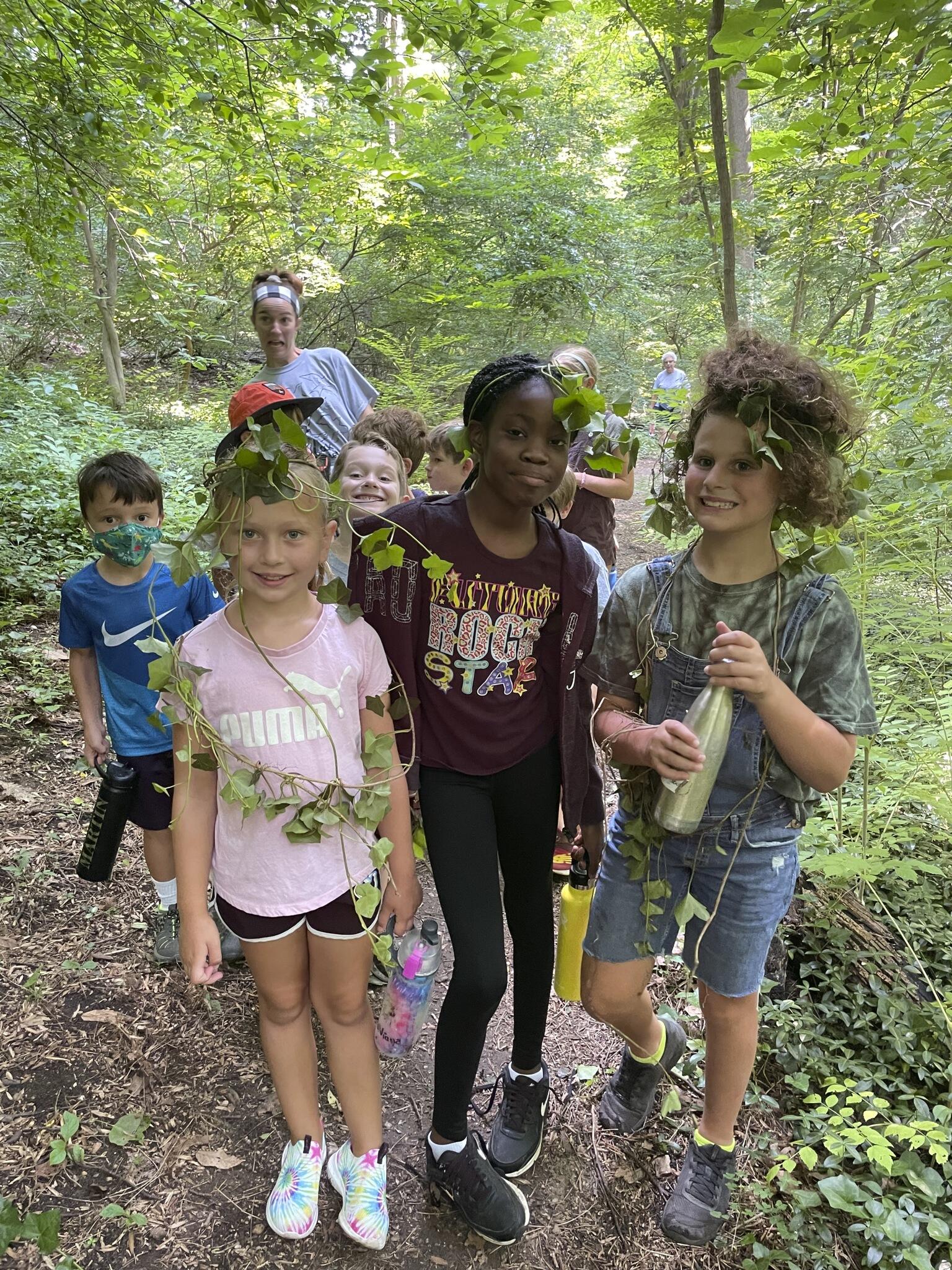 A group of children walk towards the viewer along a wooded path, smiling and wearing strands of English Ivy around them as necklaces, crowns, and general decorations.