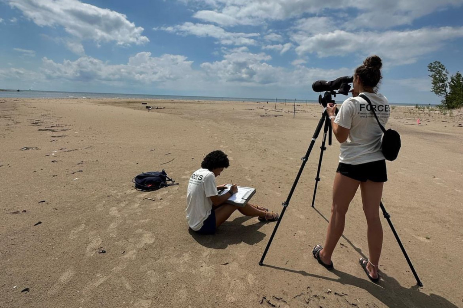 One college student sits in the sand with a clipboard, the other stands behind a scope, observing Piping Plovers