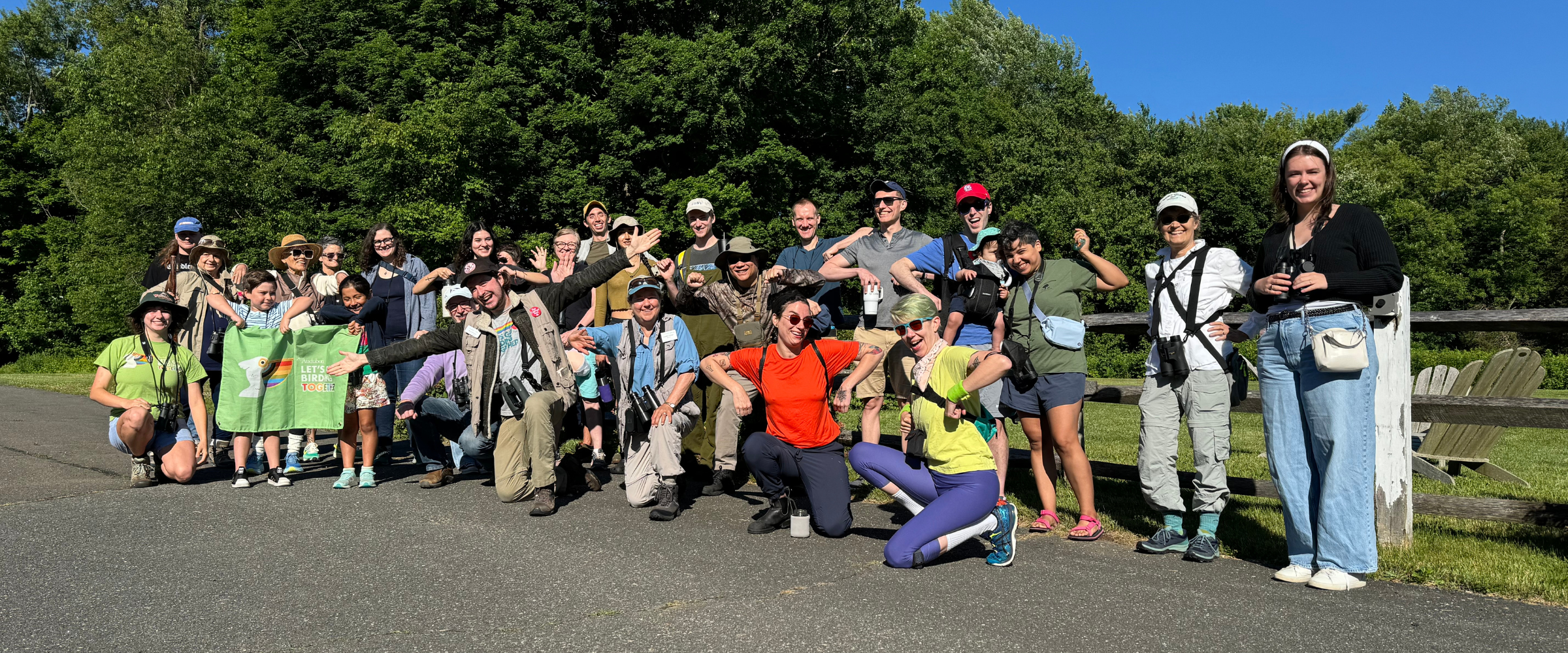 A large group of people with binoculars on a path at the Greenwich Audubon Center, posing for a photo and smiling. One person is holding up a Let's Go Birding Together banner.